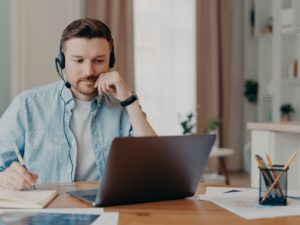 Smart man freelancer listens important info online makes necessary notes in diary works on laptop