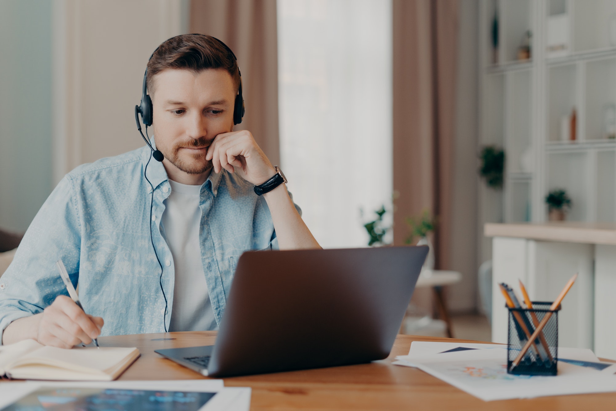 Smart man freelancer listens important info online makes necessary notes in diary works on laptop