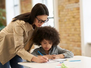 Guiding young female teacher in glasses helping little boy with the task. Kid studying in
