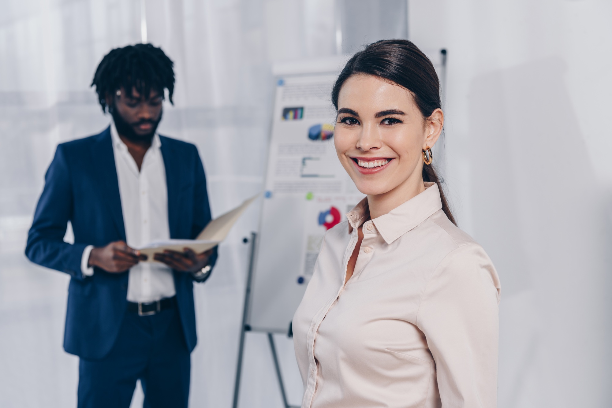 Recruiter smiling and looking at camera and african american employer near flip chart with folder in